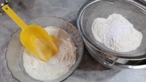 Brašno se prosejava u činiju dok se pored nalazi dodatna količina spremna za postepeno dodavanje u testo